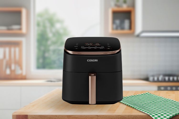 Een zwarte COSORI airfryer met een strak digitaal display siert het aanrecht. De elegante bronzen handgreep zorgt ervoor dat hij opvalt tussen de beste airfryers. Op de achtergrond maken houten planken, een potplant en een fornuis het tafereel compleet, terwijl er een groen-wit geblokte doek naast ligt.