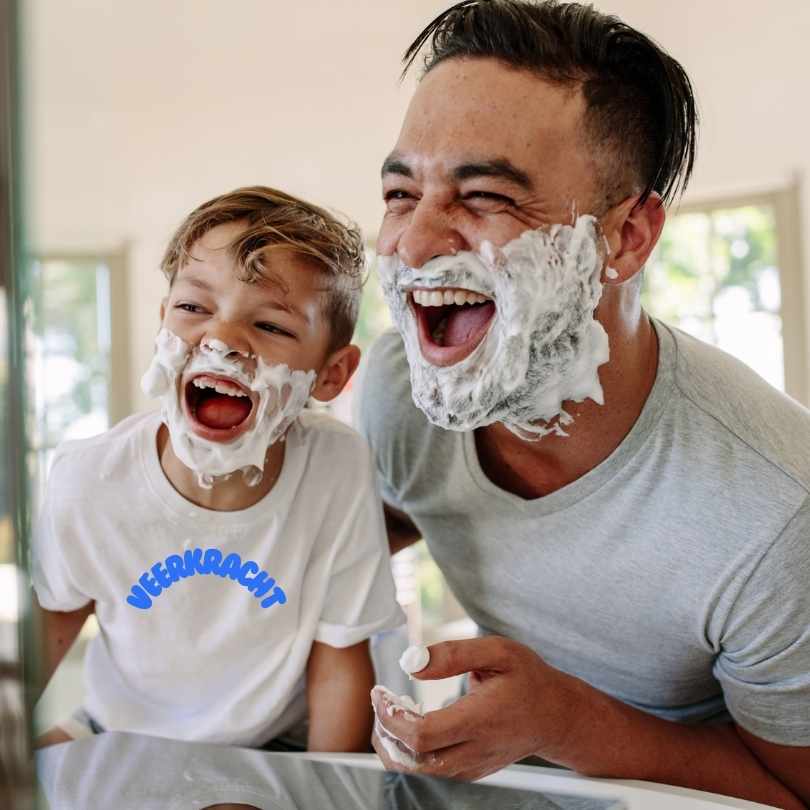 Een man en een jonge jongen, die veerkracht belichamen, lachen terwijl ze scheerschuim aanbrengen in een felverlichte badkamer. De man draagt een lichtgrijs shirt, terwijl de jongen een wit shirt met blauwe tekst draagt. Ze staan dicht bij elkaar en delen dit vreugdevolle moment.