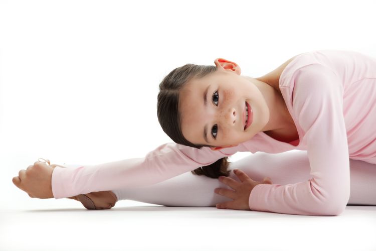 Een jong meisje in een roze balletpakje strekt zich uit op de vloer en lacht naar de camera. Haar rechterbeen is naar voren gestrekt en ze leunt eroverheen, haar voet met beide handen vastpakkend. De achtergrond is helderwit, wat haar sierlijke houding en expressie benadrukt.