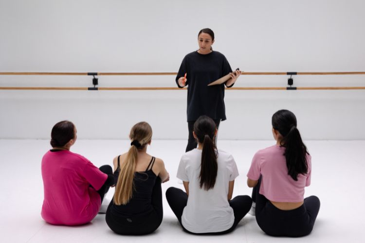 Een dansleraar in een zwarte outfit staat voor vier zittende studenten in een dansstudio met een houten barre aan de muur. De studenten, gekleed in casual sportkleding in roze, zwart en wit, luisteren aandachtig. De kamer heeft een helder, minimalistisch design.